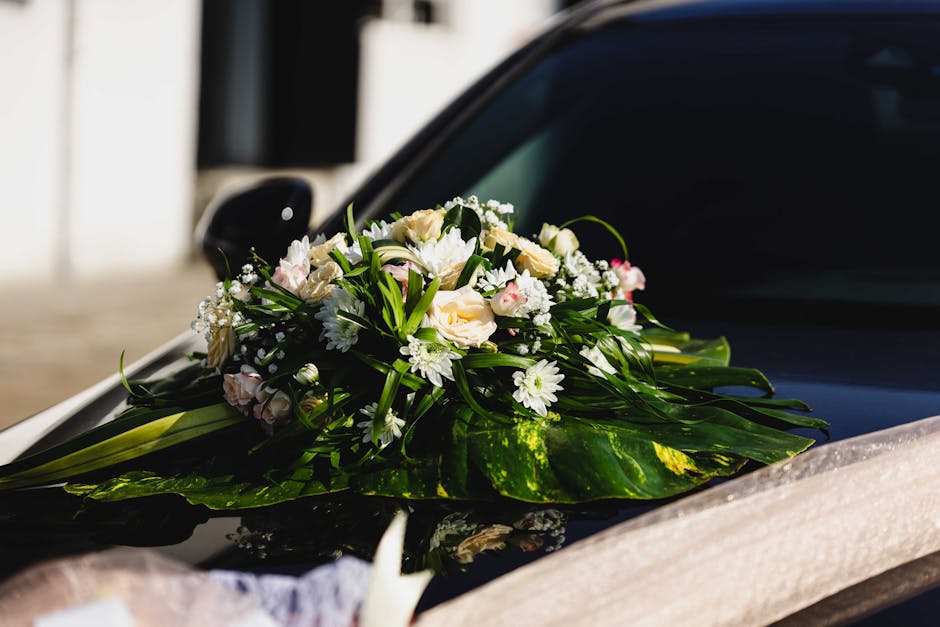 A close-up of a wedding floral arrangement resting on the hood of a black car, featuring a bouquet of fresh flowers including pale pink and white roses, white daisies, and small white baby's breath, a