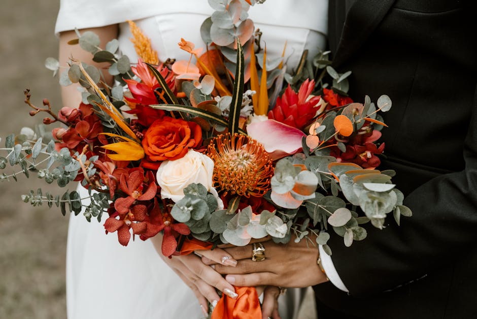 A close-up of a floral bouquet held by a person in a wedding outfit, featuring a vibrant arrangement of flowers including orange roses, white calla lilies, red protea, and pink anthuriums. Eucalyptus