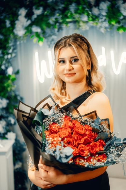 A young woman with fair skin and shoulder-length wavy blonde hair, dressed in a black sleeveless top, is holding a vibrant bouquet of fresh roses and mixed flowers wrapped in dark paper with a clear c
